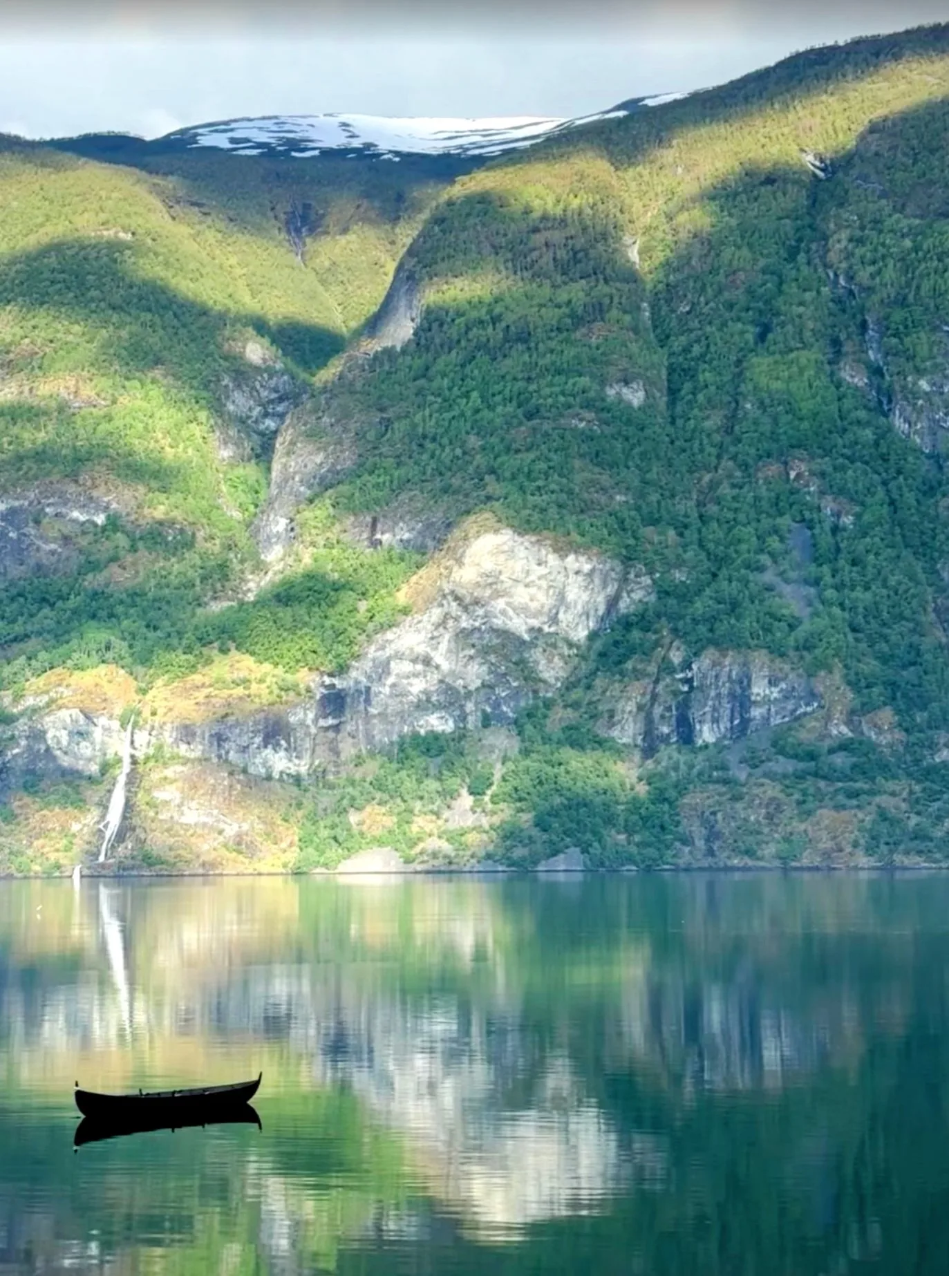 A lone boat on a Norwegian fjord, green mountains and a waterfall reflected in still water